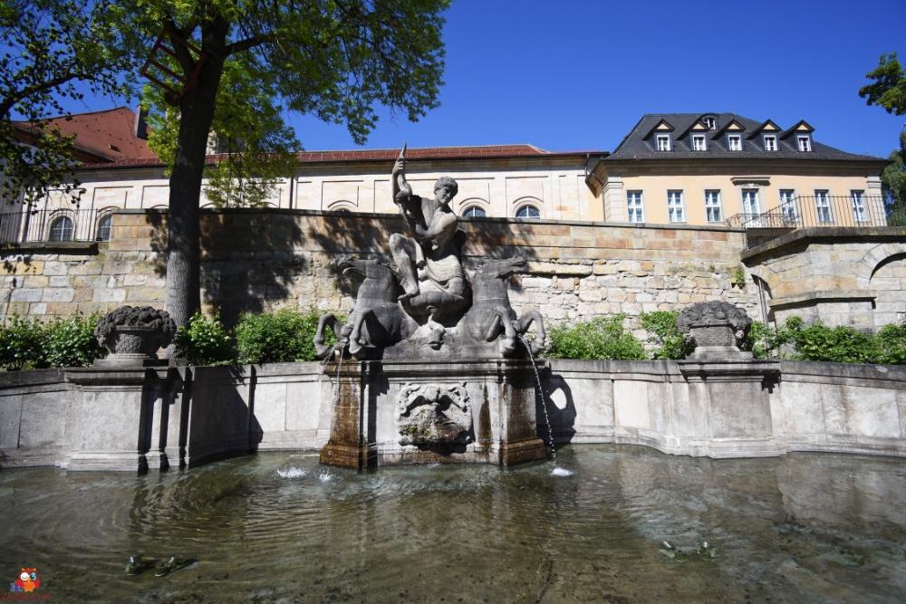 Brunnen mit einer Statue, die einen Mann darstellt, der ein Horn bläst, umgeben von weiteren Figuren, Grünanlagen und historischen Gebäuden im Hintergrund.