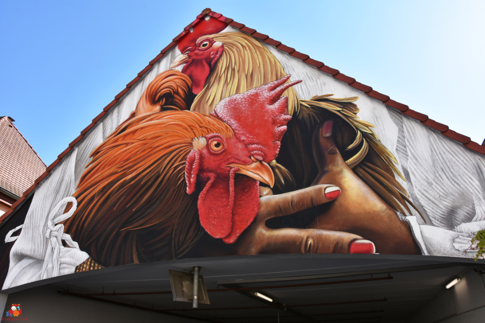 Großes Wandgemälde eines Huhns und einer Hand mit bunten Nägeln, das auf einem Dach zu sehen ist.