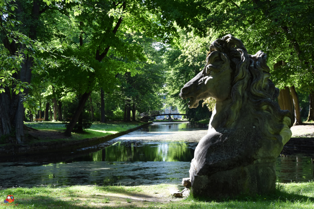 Skulptur eines Pferdekopfes aus Stein im Hofgarten von Bayreuth, umgeben von üppigem Grün und einem ruhigen Kanal im Hintergrund.