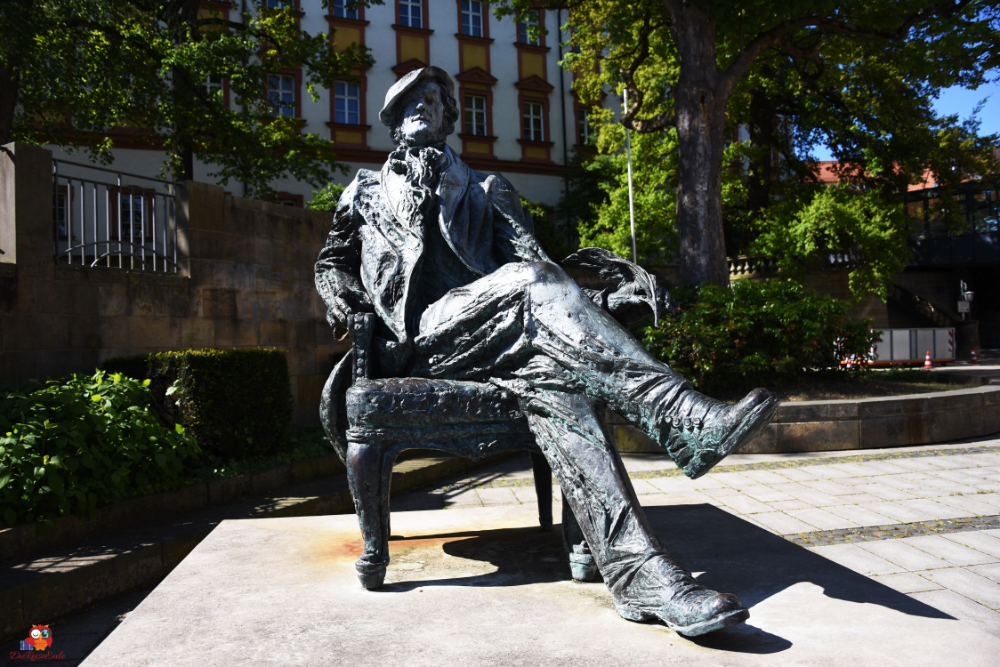 Bronzestatue von Richard Wagner, sitzend auf einer Bank, umgeben von grünen Pflanzen und historischem Gebäude im Hintergrund.