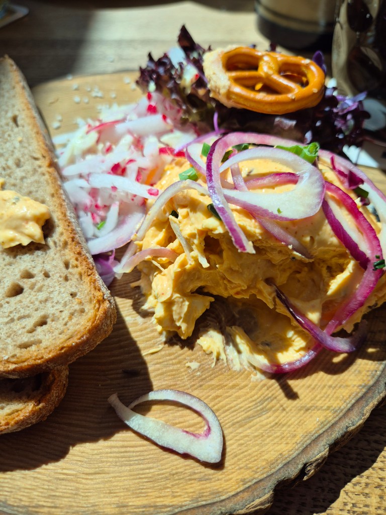 Ein Teller mit Brot, frischem Salat, Zwiebeln und einer cremigen Aufstrichsorte, garniert mit einer Brezel.