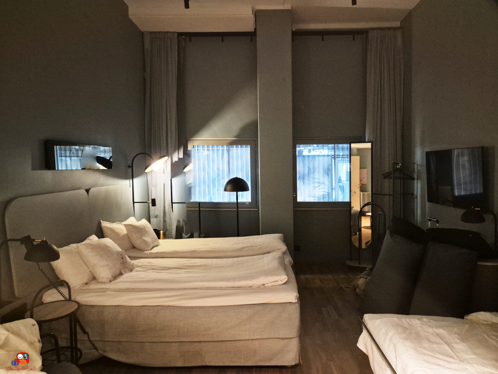 Ein modernes Hotelzimmer mit zwei Einzelbetten und einer Ausziehcouch, ausgestattet mit Leselampen und einem großen Fernseher, sowie zwei Fenstern, die auf eine Baustelle blicken.