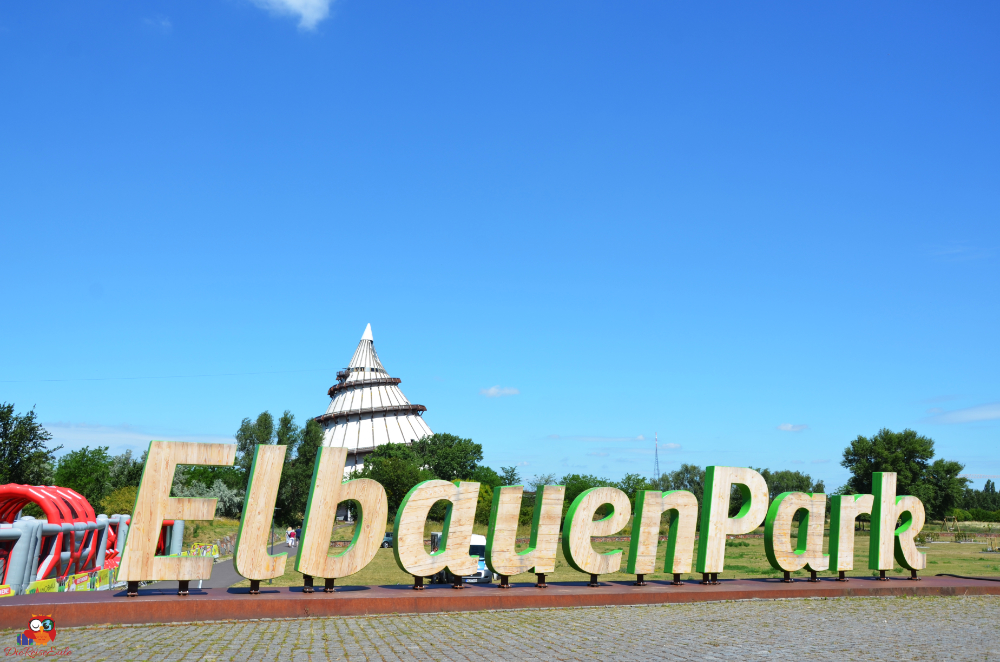 Schriftzug 'ElbauenPark' auf einem Platz, im Hintergrund ein hoher, hölzerner Turm und blauer Himmel.