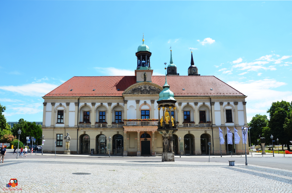 Das Alte Rathaus in Magdeburg mit seiner beeindruckenden Architektur und den charakteristischen Türmen, umgeben von einem Platz mit Bäumen und Flaggen.