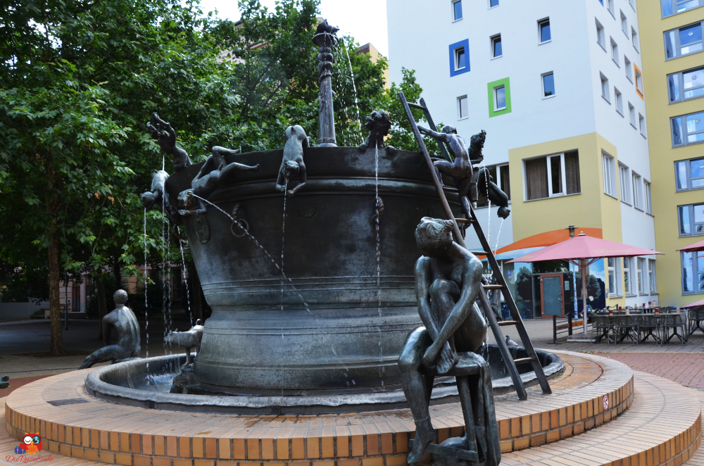 Brunnen mit Skulpturen von menschlichen Figuren, die um eine zentrale Wasserstelle sitzen und klettern, umgeben von Bäumen und modernen Gebäuden in Magdeburg.
