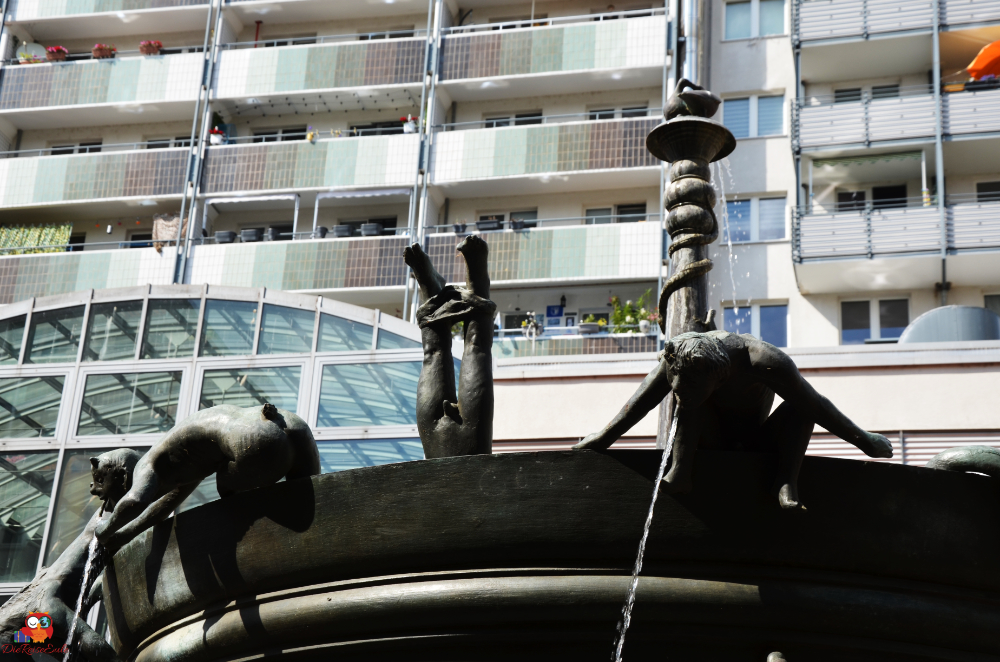 Brunnen mit skulpturalen Figuren, die aus der Mitte strömen, im Vordergrund; im Hintergrund moderne Wohngebäude.
