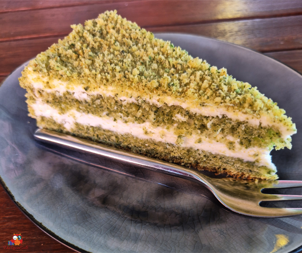 Ein Stück Kuchen auf einem Teller mit einer Gabel daneben. Der Kuchen hat eine grüne Farbe mit weißen Schichten und einer krümeligen Oberfläche.