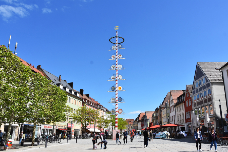 Blick auf einen Platz in einer Stadt mit einem hohen Maibaum und verschiedenen Geschäften und Cafés im Hintergrund.