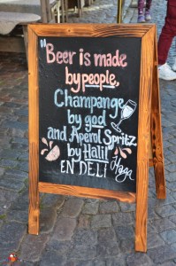 Schwarze Tafel mit handschriftlichem Spruch über Bier, Champagner und Aperol Spritz, dekorativ umrahmt und auf einem Holzständer auf einem gepflasterten Platz aufgestellt.