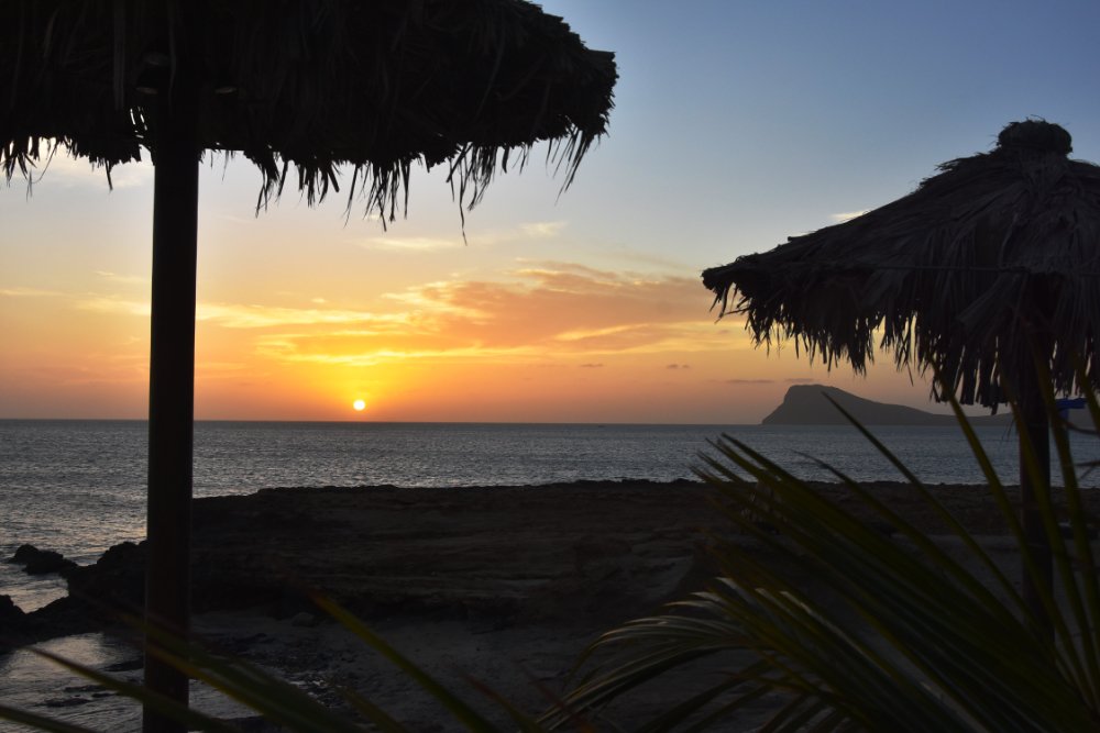 Sonnenuntergang am Strand mit Palmen und Strandzelt im Vordergrund.