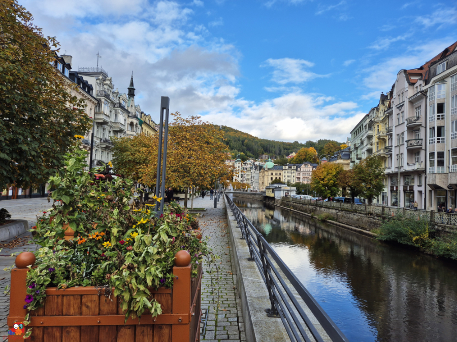 Blick auf eine malerische Stadt mit bunten Gebäuden und einem ruhigen Fluss, umgeben von herbstlichen Bäumen und einem blauen Himmel.