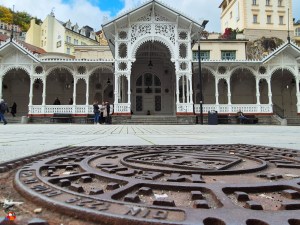Detailaufnahme einer Gussdeckel mit einem historischen Motiv im Vordergrund, während im Hintergrund ein elegantes Gebäude mit weißem Holzfachwerk und Bogenfenstern zu sehen ist.