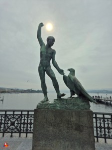 Bronzeskulptur eines nackten Jungen mit einem Adler, der seine Hand ausstreckt. Der Junge steht auf einem Podest mit der Inschrift "Ganymed", im Hintergrund ein See und ein bewölkter Himmel.