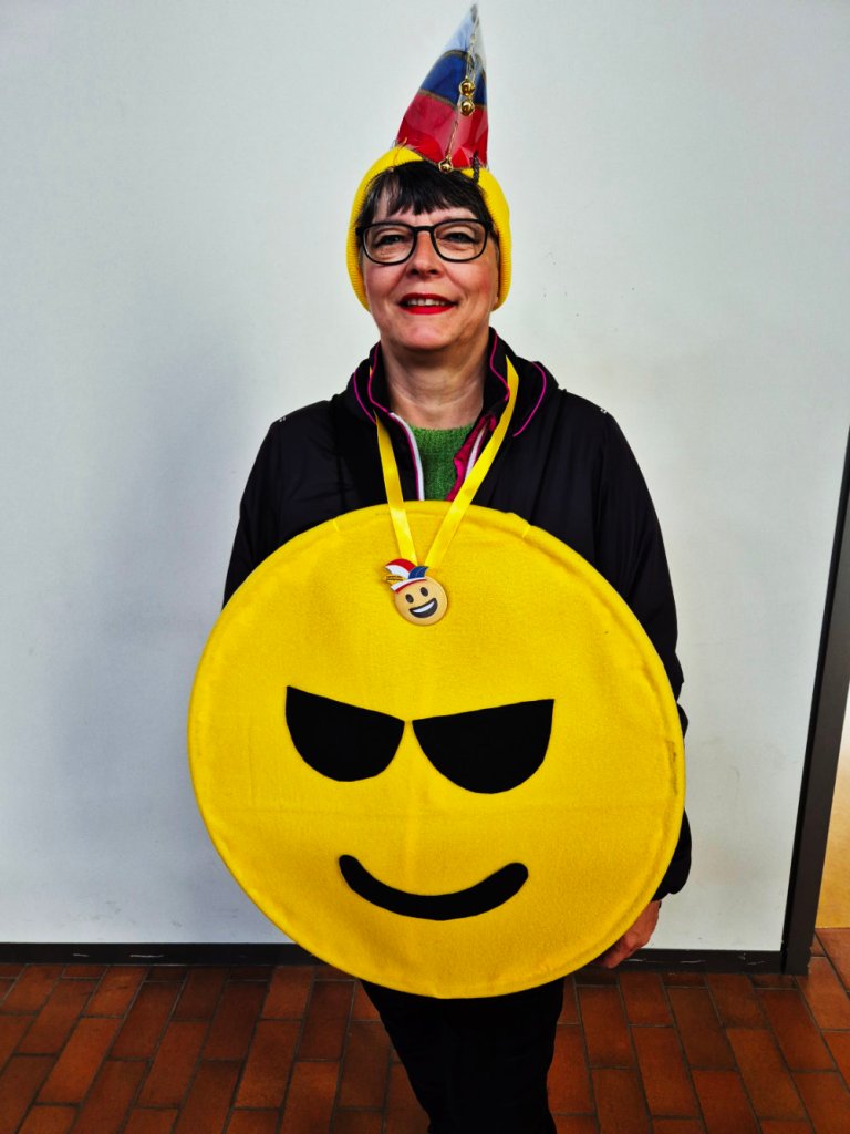 Eine Frau in einem gelben Smiley-Kostüm mit Sonnenbrille, einer bunten Hut und einer Medaille, steht vor einer weißen Wand.