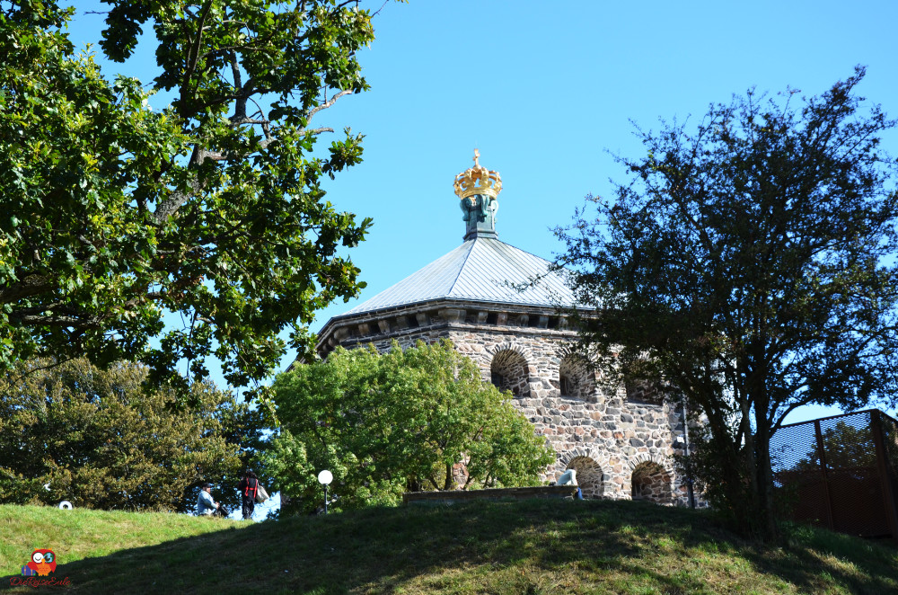 Eine historische Steinfabrik mit einem markanten Dach und einer goldenen Krone, umgeben von Bäumen und grünem Rasen.
