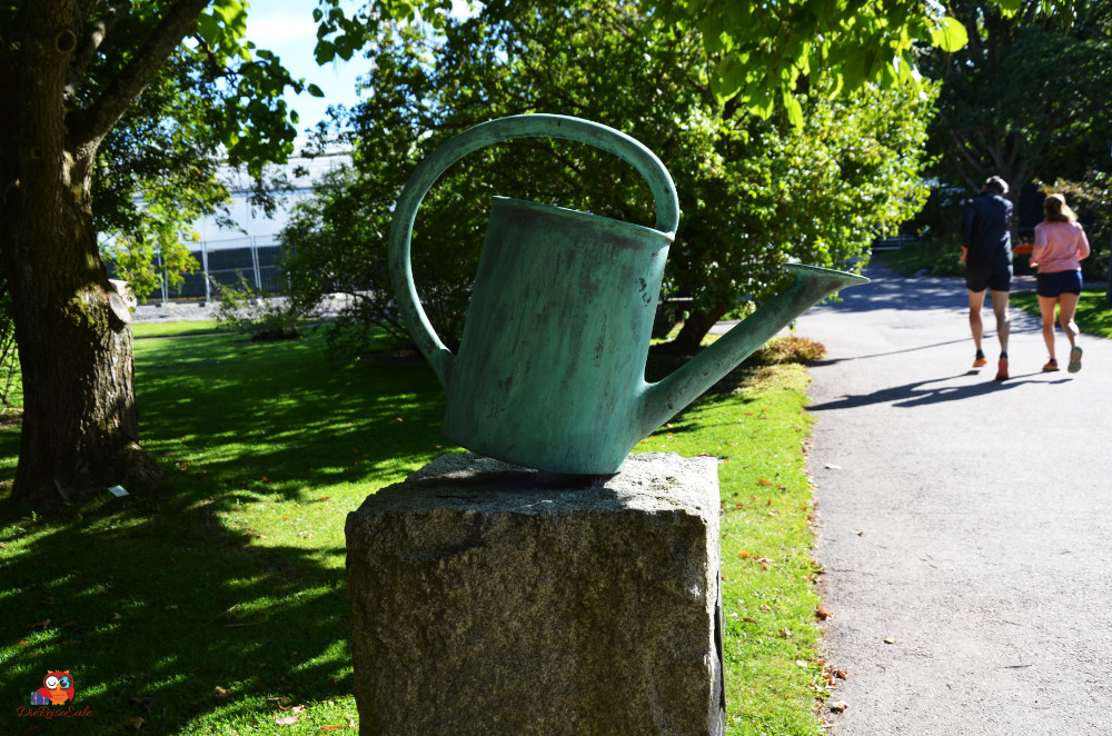 Ein bronzegrüner, stilisierter Gießkanne aus Metall, die auf einem Steinuntergrund steht, im Hintergrund sind Spaziergänger in einem parkähnlichen Bereich zu sehen.
