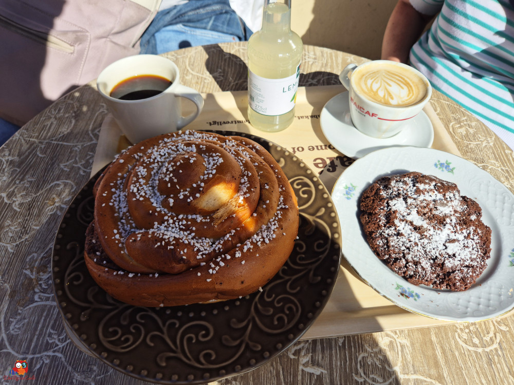 Ein Teller mit einer großen Zimtschnecke, einem Keks und Bechern mit Kaffee und einem Getränk auf einem Tisch.