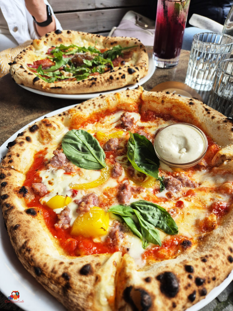 Ein Teller Pizza mit einem dicken, knusprigen Rand, belegt mit Wurst, gelben Paprikastücken, Basilikum und einem Dip auf der Seite. Im Hintergrund ist eine weitere Pizza und Getränke zu sehen.