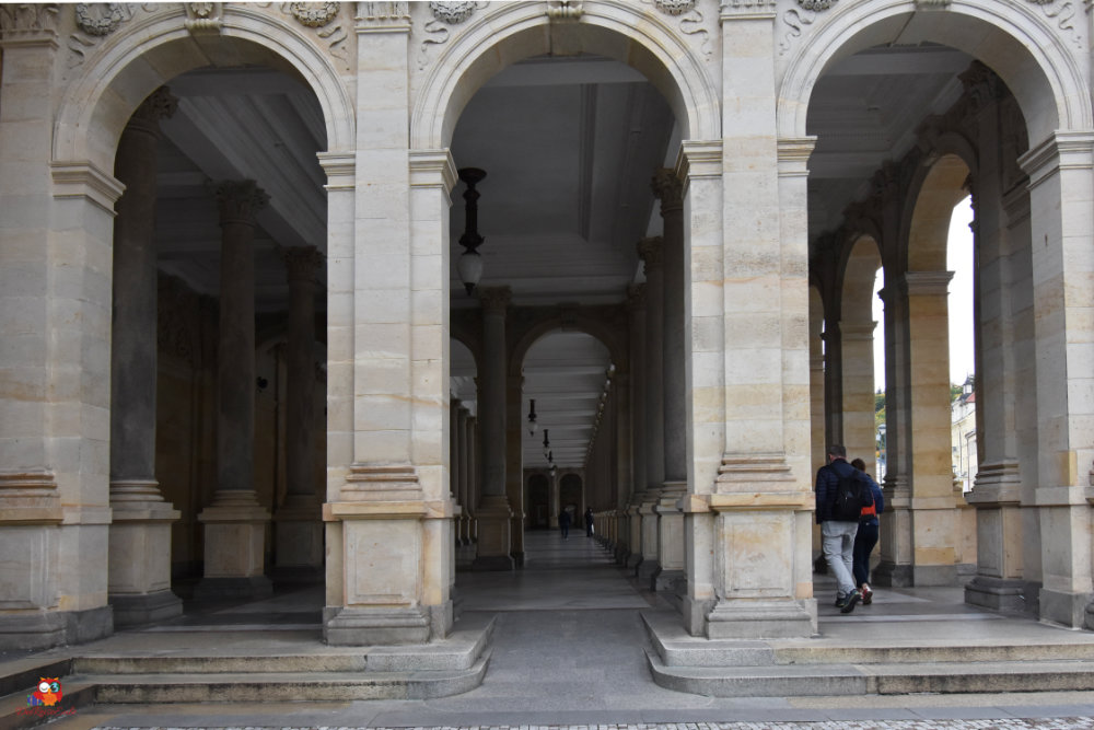 Ein Säulengang mit mehreren Bögen und Säulen, in dem zwei Personen entlanggehen.