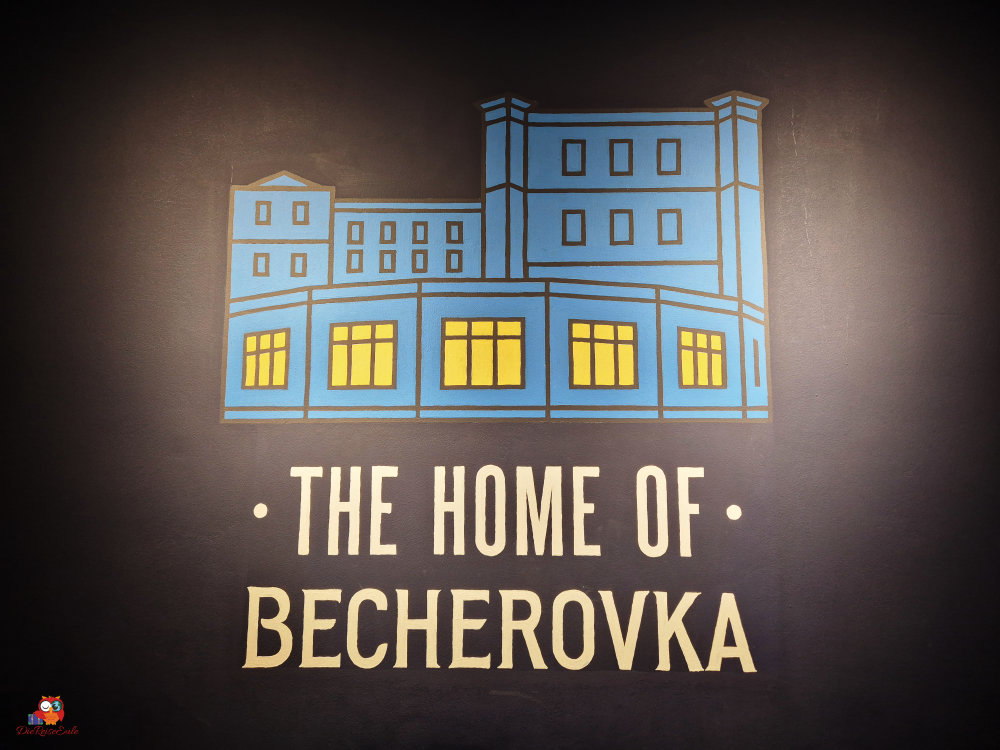 logo von becherovka, skizze des gebäudes in blau mit gelben fenstern auf schwarzem grund, darunter die schrift: the home of becherovka