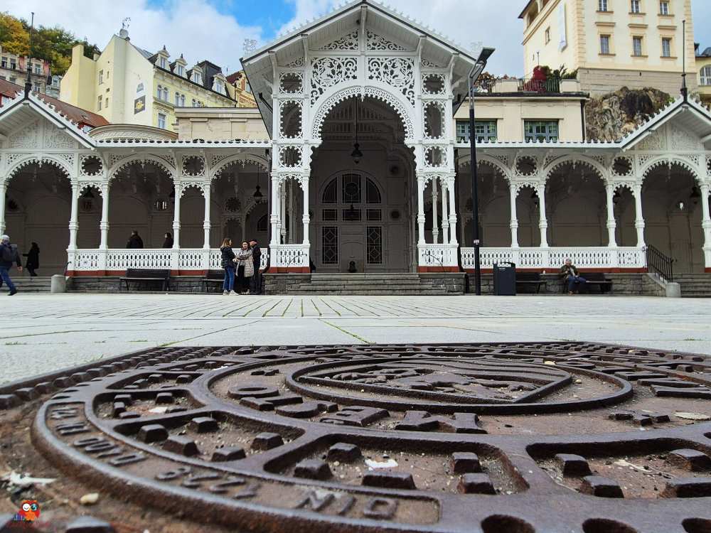 Historisches Gebäude mit verzierter Holzveranda in einem Park, im Vordergrund ein Menchlochdeckel.