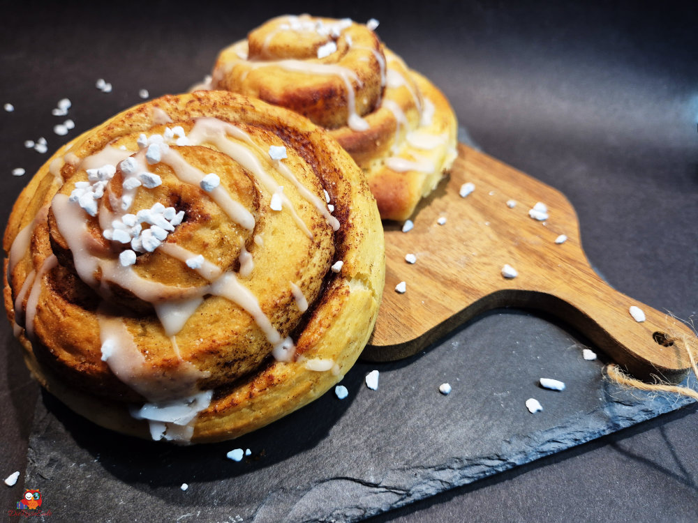 Zwei frisch gebackene Zimtschnecken mit Zuckerguss und Perlzucker auf einem Holzbrett, dekoriert auf einem schwarzen Hintergrund.