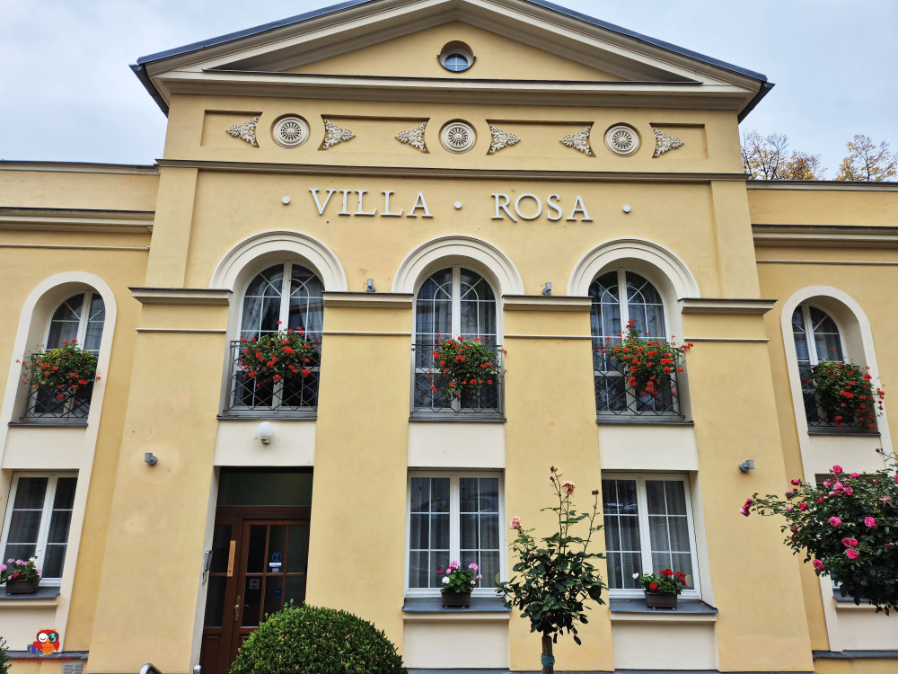 Ein einfaches Schlafzimmer mit zwei Bettgestellen, die mit weißen Bettdecken und Kissen ausgestattet sind. An der Wand hängt ein Fernseher, und es gibt einige Bilder an der Wand. Die Fenster sind mit roten Vorhängen dekoriert.