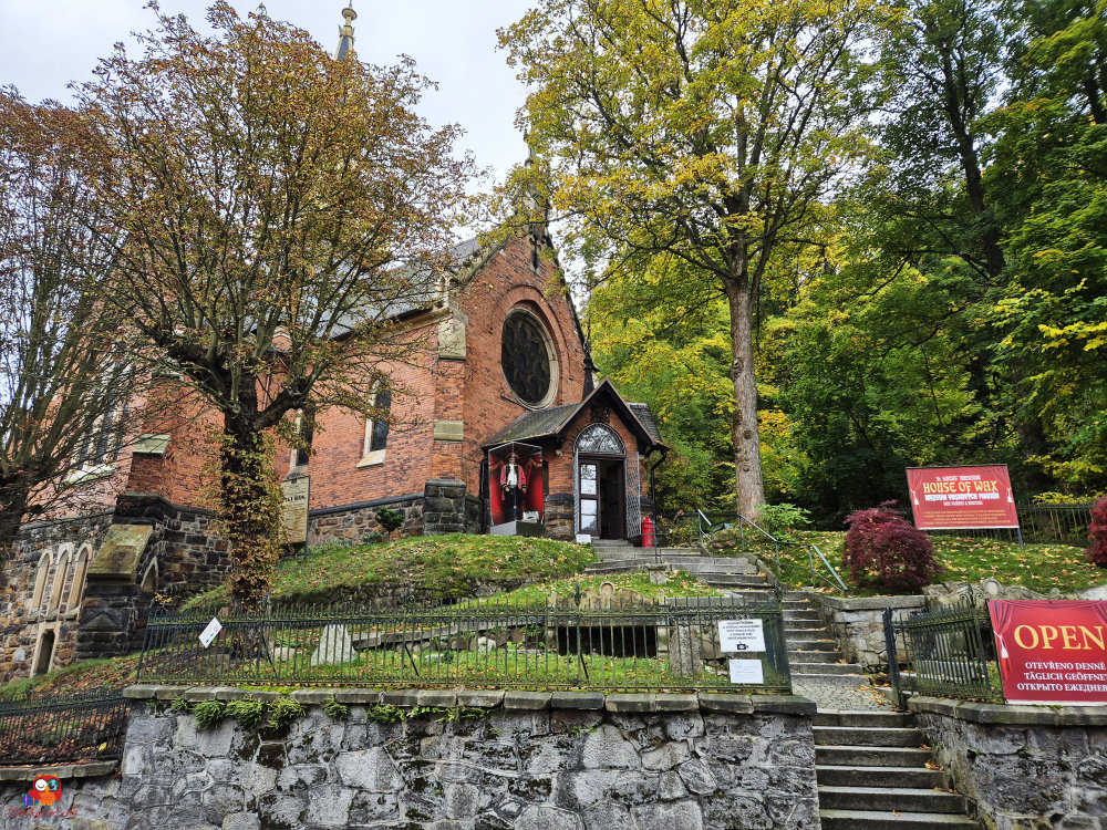 Eine historische Gebäudeansicht mit roten Ziegeln, umgeben von herbstlichen Bäumen. Treppe führt zu einem Eingang mit einem Schild 'OPEN' und einem weiteren Schild 'HOUSE OF WAX'.
