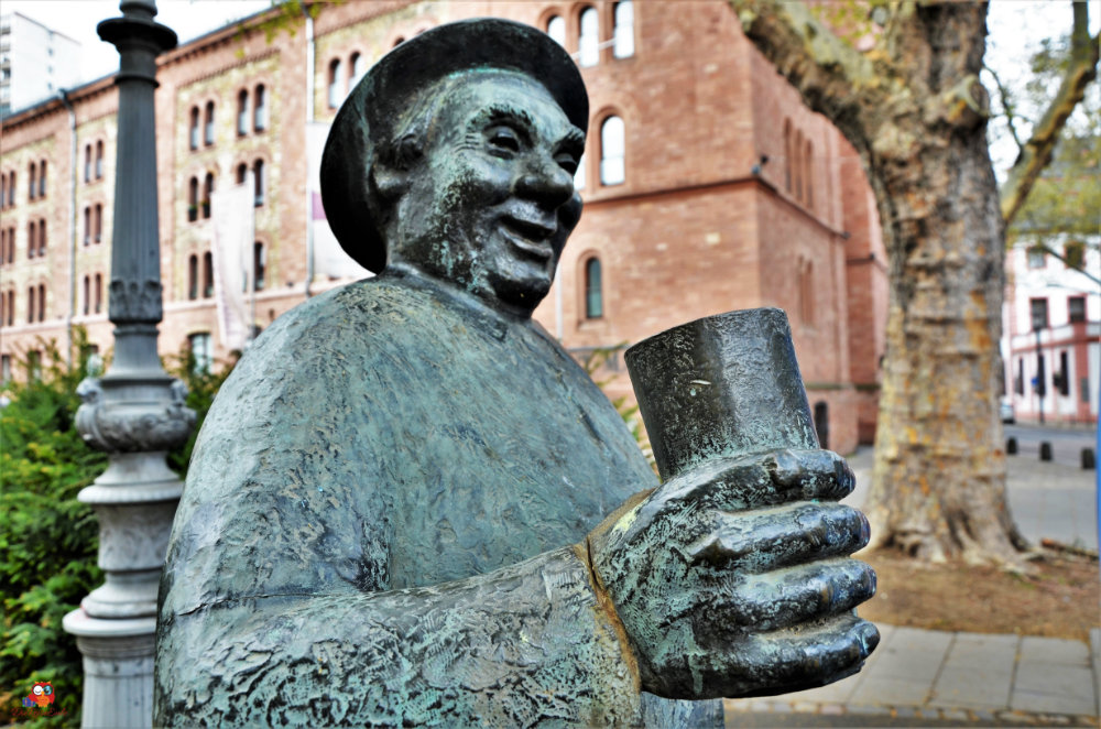 Bronzestatue eines lächelnden Mannes, der einen Becher hält, im Vordergrund. Im Hintergrund ist ein historisches Gebäude und Bäume zu sehen.