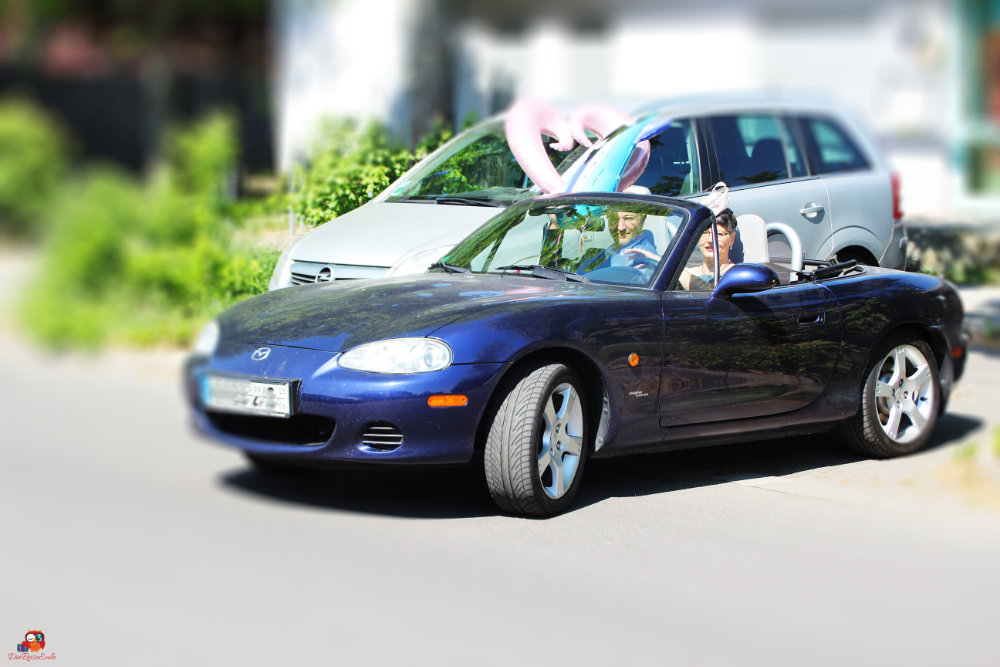 Blauer Cabrio-Sportwagen mit zwei Personen im Inneren undließen Luftballons in Herzform, geparkt an einer Straße.