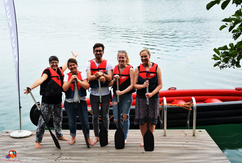 Fünf Personen in Schwimmwesten stehen am Ufer eines Gewässers mit Paddeln in der Hand, im Hintergrund ist ein aufblasbares Boot zu sehen.