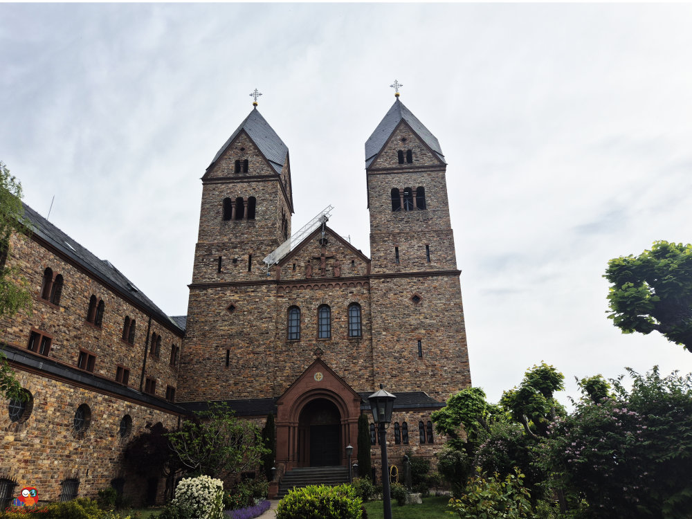 Das Bild zeigt eine historische Kirche mit zwei Türmen, die aus Stein gebaut ist, umgeben von einem grünen Garten und einem bewölkten Himmel.