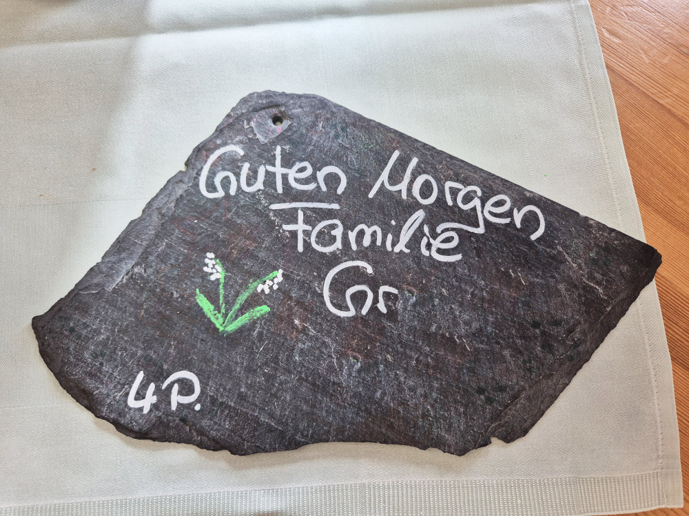 Eine schwarze Steinplatte mit der Aufschrift 'Guten Morgen Familie G.' und einer handgemalten Blume.