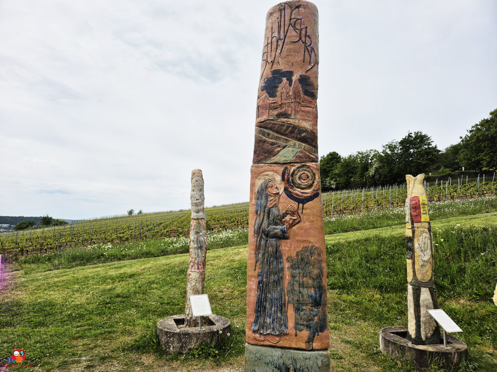 Drei farbige Skulpturen in einem Weinberg, mit einer großen Säule in der Mitte, die eine Frau und verschiedene Motive darstellt.