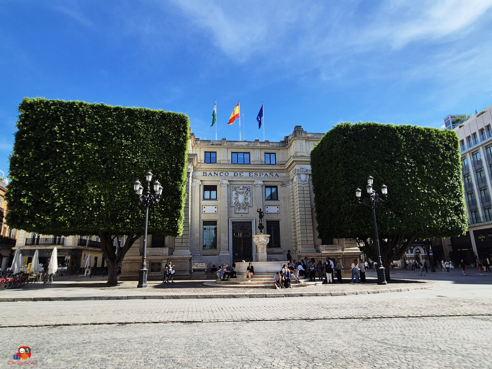 Das Gebäude des Banco de España mit großflächigen, grünen Hecken und Flaggen, umgeben von Menschen und Cafés auf dem Platz.