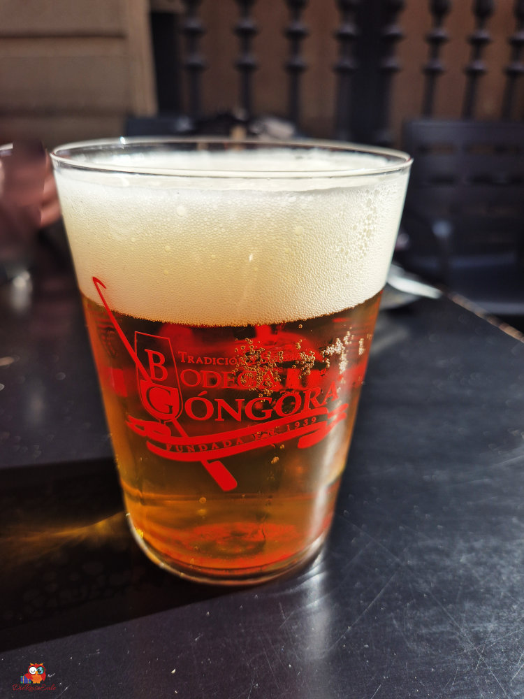 Ein Glas Bier mit Schaum, bedruckt mit dem Logo von Bodega Góngora, auf einem Tisch.