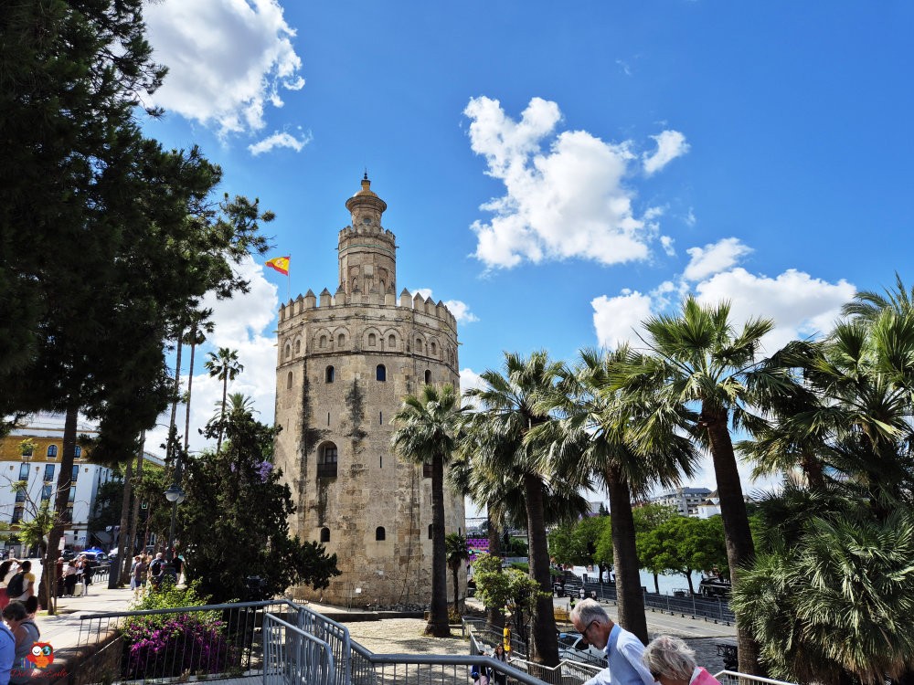 Ein historischer Turm mit einer charakteristischen Spitze, umgeben von Palmen und einem blauen Himmel mit Wolken.