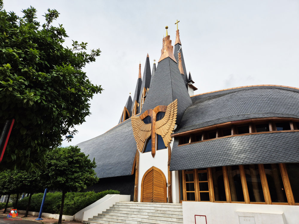 Ansicht einer modernen Kirche mit markanten, spitzen Türmen und einer hölzernen Fassade, die eine stilisierte Vogelgestaltung zeigt.