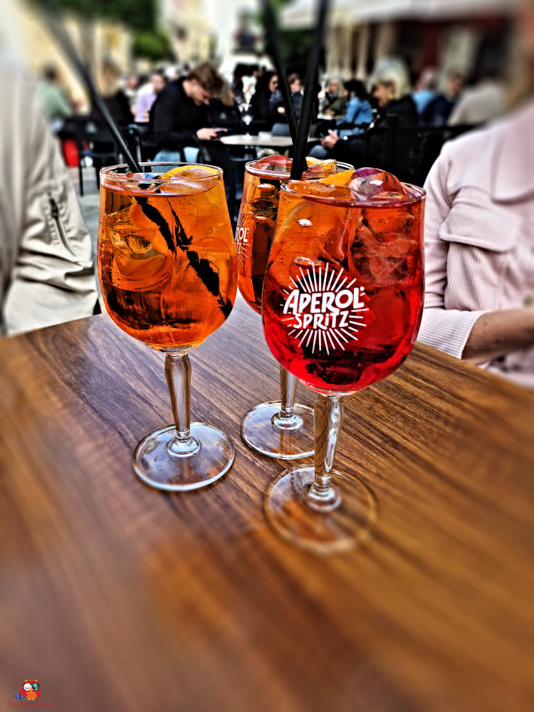 Drei Aperol Spritz Getränke in großen Gläsern mit Eis und Trinkhalmen auf einem Holztisch.